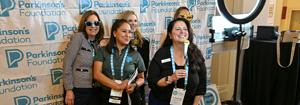 Five smiling women posing in front of a Parkinson's Foundation backdrop with event badges and props, next to a ring light.