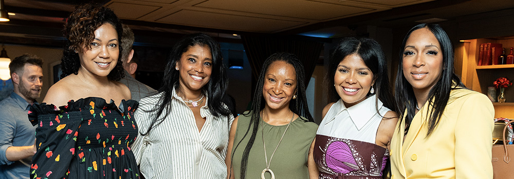 Group of five diverse women smiling indoors at a social event or gathering.