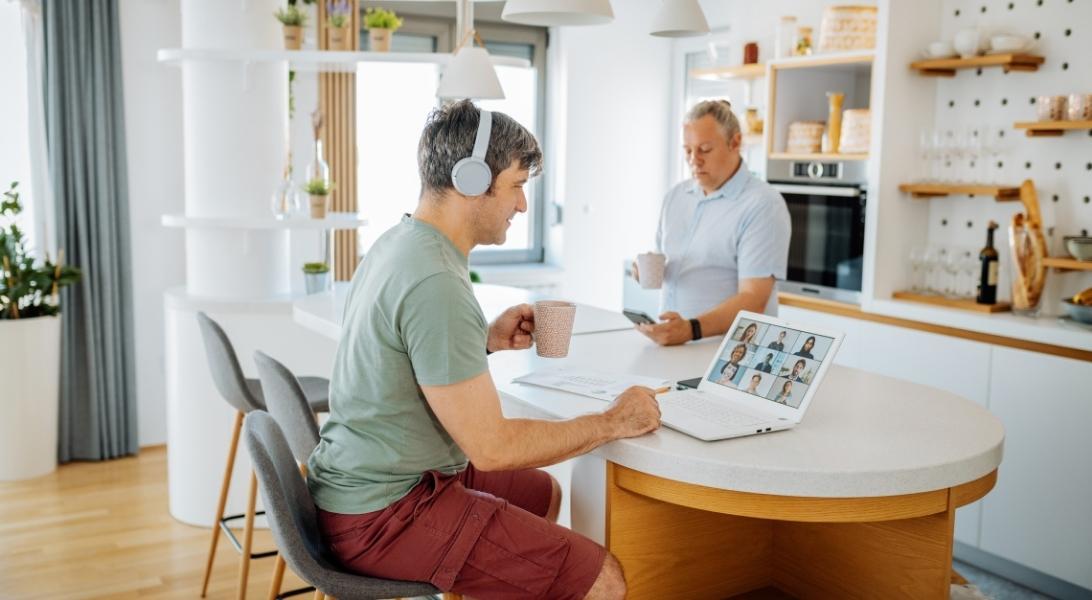 Man sitting at counter with headphones in and coffee mug in hand, watching a Zoom call on his laptop. Another man stands in background, with coffee mug in one hand and smartphone in the other.