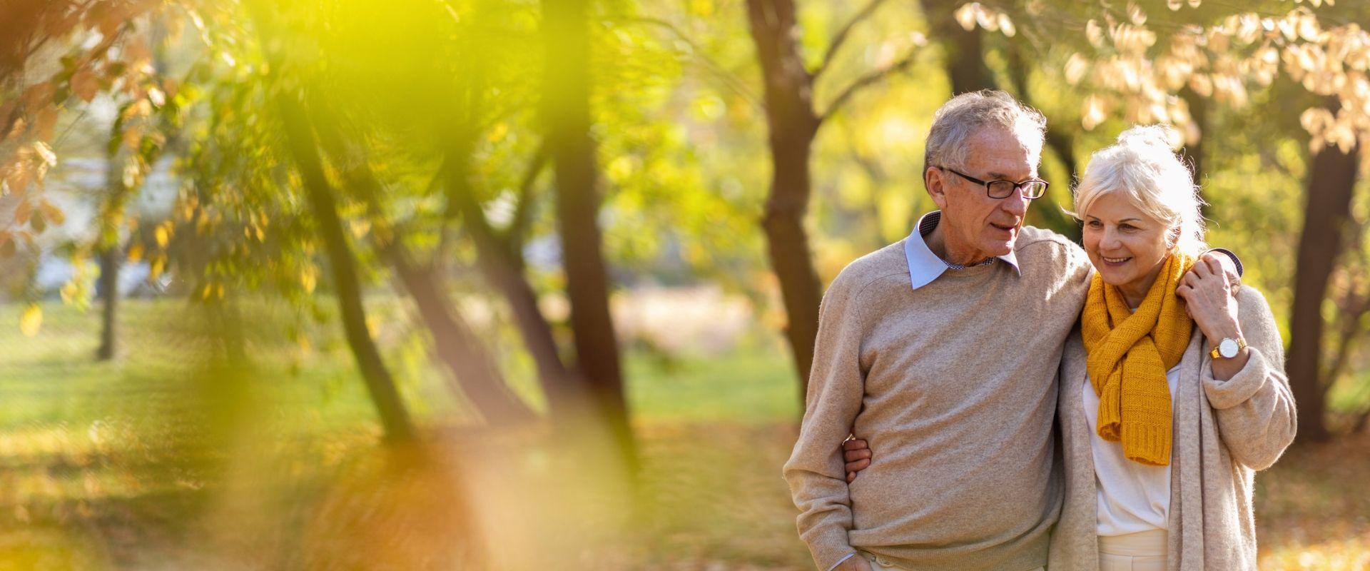 Older couple walking arm in arm through a sunlit park in fall.