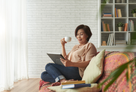 An older asian woman sitting on a couch, reading, drinking from a mug.