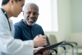 A black man in a dark blue shirt speaks to an Asian doctor in a waiting room, who is showing him something on a clipboard.