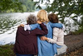 Three women sitting on a bench by a lake hugging