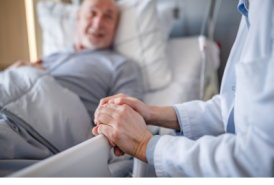 A senior man lies in a hospital bed holding hands with a doctor.