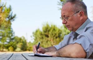 Man sitting at picnic table writing in notebook with a pen