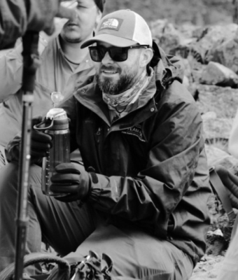 Black and white photo of a bearded man wearing sunglasses, a cap, and outdoor gear, sitting on a rocky surface and holding a water bottle.