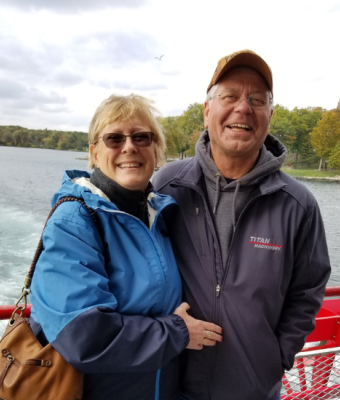 Smiling couple in jackets standing on a boat with a lake and trees in the background.