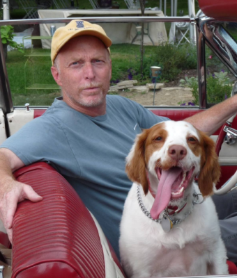 A man in a yellow cap and blue shirt sits in a red vintage car with a happy brown-and-white dog beside him.