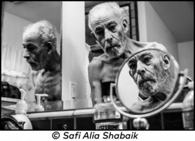 Black and white portrait of Aly H. Shabaik, sitting in a chair and looking at his shirtless reflection in a mirror