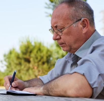 Man sitting at picnic table writing in notebook with a pen 