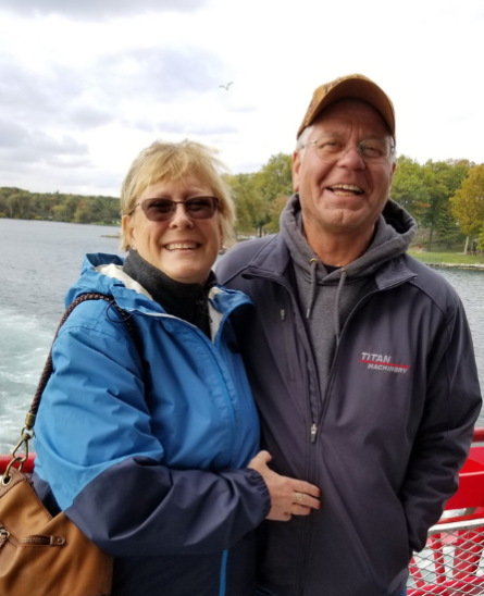 Smiling couple in jackets standing on a boat with a lake and trees in the background.