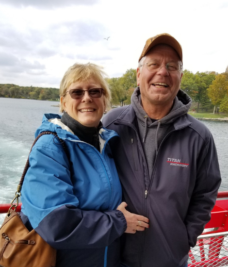 Smiling couple in jackets standing on a boat with a lake and trees in the background.