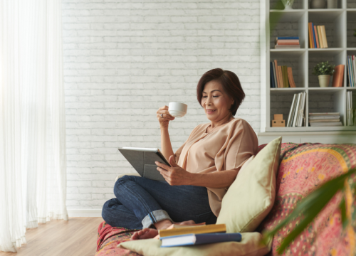 An older asian woman sitting on a couch, reading, drinking from a mug.