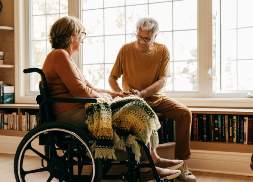 A woman in a wheelchair sits in front of a wall of windows as a man tenderly cares for her