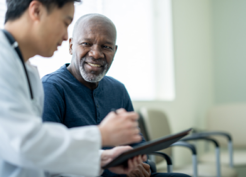 A black man in a dark blue shirt speaks to an Asian doctor in a waiting room, who is showing him something on a clipboard.