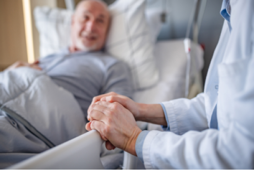 A senior man lies in a hospital bed holding hands with a doctor.