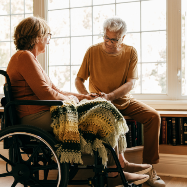A woman in a wheelchair sits in front of a wall of windows as a man tenderly cares for her