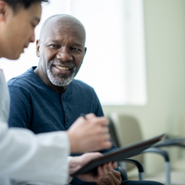 A black man in a dark blue shirt speaks to an Asian doctor in a waiting room, who is showing him something on a clipboard.
