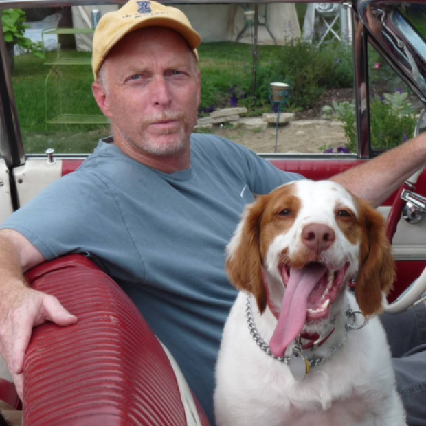 A man in a yellow cap and blue shirt sits in a red vintage car with a happy brown-and-white dog beside him.
