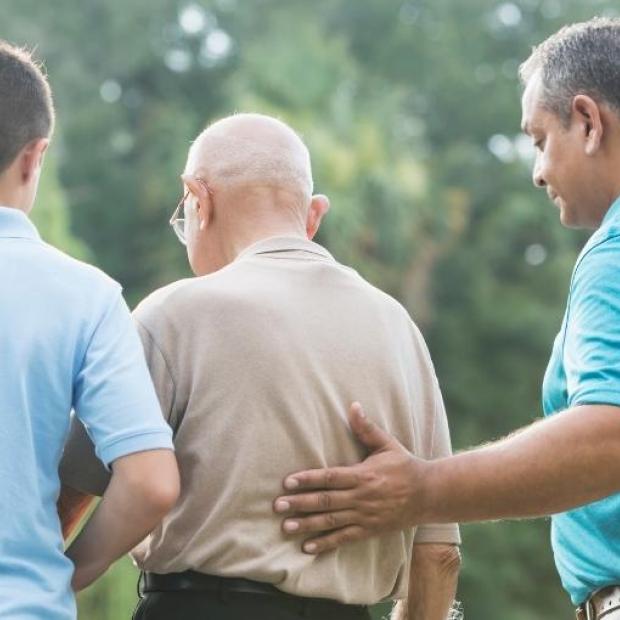 Multigenerational family of men with their backs to the camera. Grandfather stands in the middle with his grandson on the left and his son on the right. Son's left hand is placed on his father's back