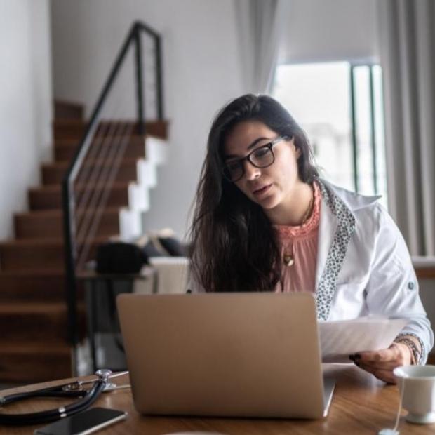 Woman sitting in chair at dining room table with papers in hand, looking at her laptop. A stethoscope, smartphone and cup of tea are placed on the table in front of her