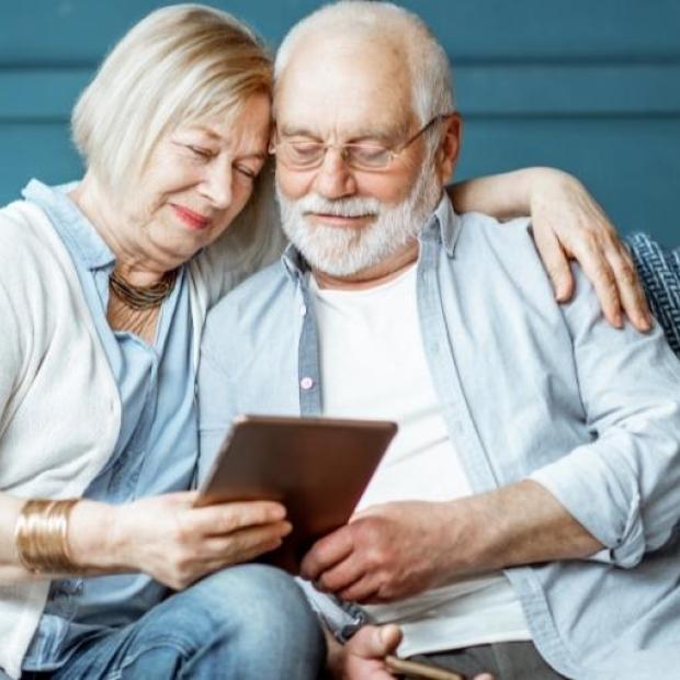 Husband and wife sitting together on the couch, looking at an iPad. Wife has her left arm over her husband's shoulder and both look happy