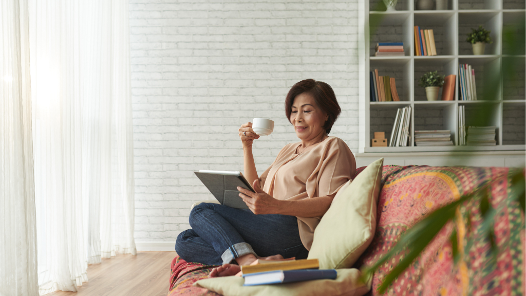 An older asian woman sitting on a couch, reading, drinking from a mug.