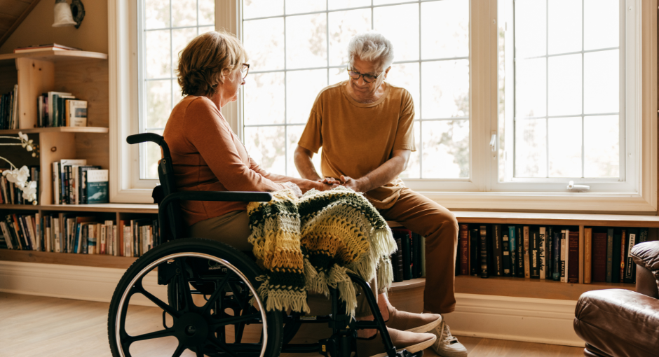 A woman in a wheelchair sits in front of a wall of windows as a man tenderly cares for her