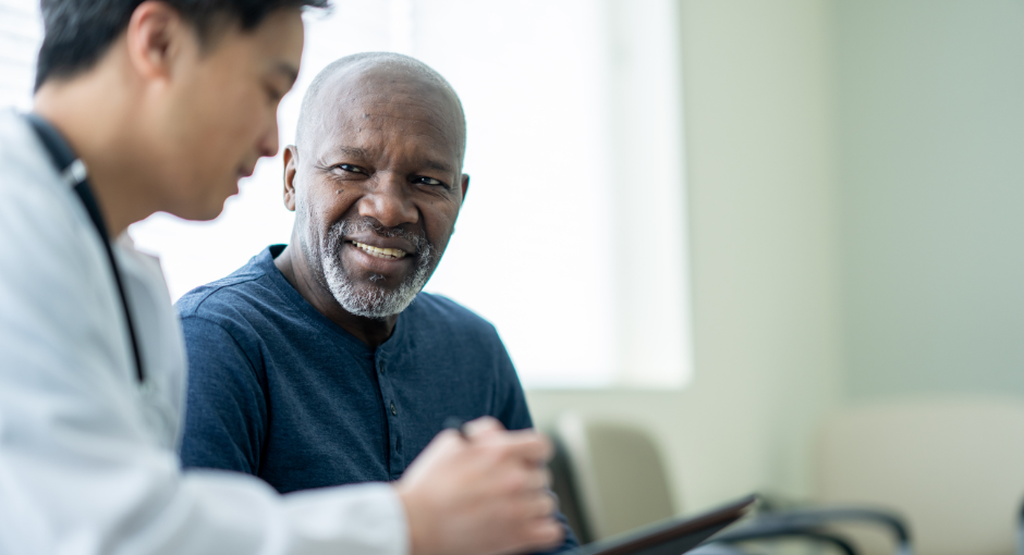 A black man in a dark blue shirt speaks to an Asian doctor in a waiting room, who is showing him something on a clipboard.