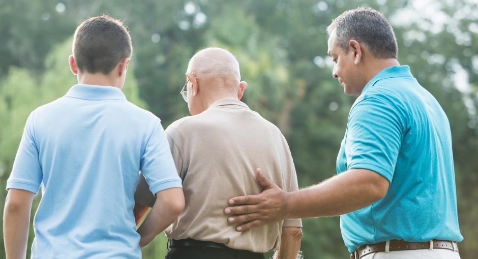 Família multigeracional de homens de costas para a câmera.  O avô fica no meio com o neto à esquerda e o filho à direita.  A mão esquerda do filho é colocada nas costas do pai