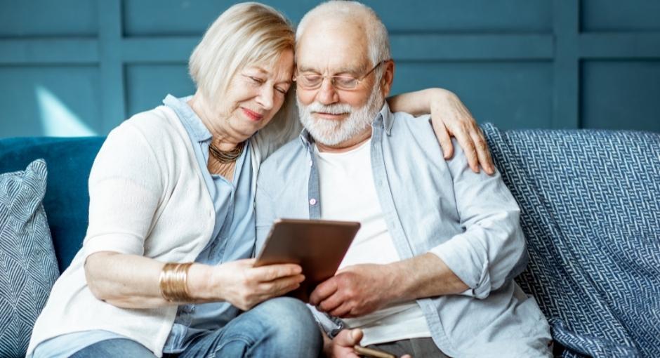 Husband and wife sitting together on the couch, looking at an iPad. Wife has her left arm over her husband's shoulder and both look happy
