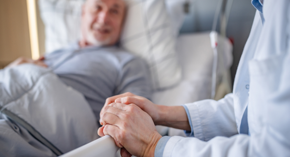 A senior man lies in a hospital bed holding hands with a doctor.