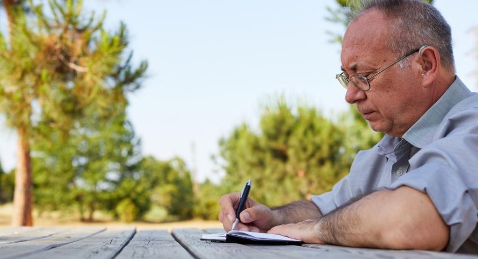 Man sitting at picnic table writing in notebook with a pen 
